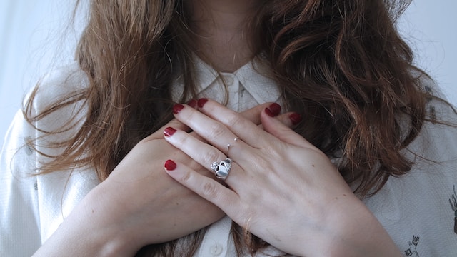 A person holding their hand to their chest. They are wearing a ring with a heart shape engraved on it.