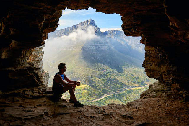 A man sitting at the mouth of a cave opening, which shows a gorgeous mountainside and fields below. 